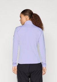 Svetlofialová dlhá rukávová športová blúza s vysokým golierom, priliehavý strih a hladká textúra. Čierne nohavice s bielymi bočnými pruhmi.