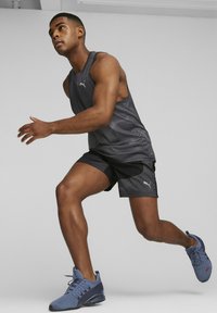 Camiseta sin mangas de entrenamiento en negro y gris con un patrón de puntos, combinada con pantalones cortos negros. El corredor lleva zapatillas deportivas azules con parte superior de malla y un diseño deportivo.