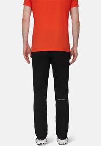 Man som står vänd bort med ryggen mot kameran, iklädd en röd kortärmad skjorta, svarta Mammut-byxor och vita träningsskor mot en enfärgad bakgrund.