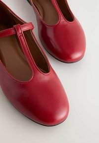 Zapatos Mary Jane rojos brillantes con una correa sobre el empeine, con punta redondeada y una superficie lisa. Situados sobre un fondo claro.
