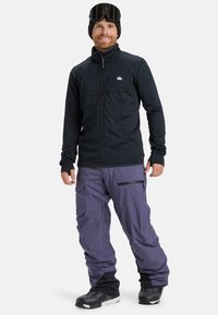 Hombre con chaqueta negra con cremallera, pantalones de nieve morados, botas de nieve negras, gorro negro y gafas de esquí en la cabeza, de pie y sonriendo.