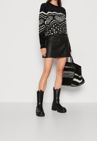 Schwarzer strukturierter Pullover mit weißen Mustern, kombiniert mit einem schwarzen Leder-Minirock. Schwarze klobige Ankle-Boots und eine gemusterte Tragetasche.