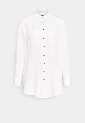 Longue chemise blanche à boutons avec manches longues, deux poches poitrine et boutons marron sur le devant, sur fond gris clair.