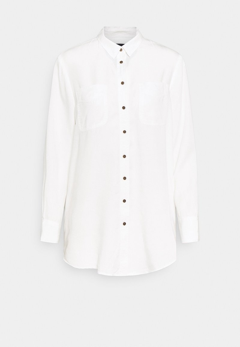 Longue chemise blanche à boutons avec manches longues, deux poches poitrine et boutons marron sur le devant, sur fond gris clair.