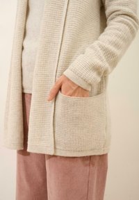 Cárdigan de punto beige claro con un acabado texturizado, con apertura frontal y bolsillos laterales. Combinado con pantalones de pana rosa suaves.