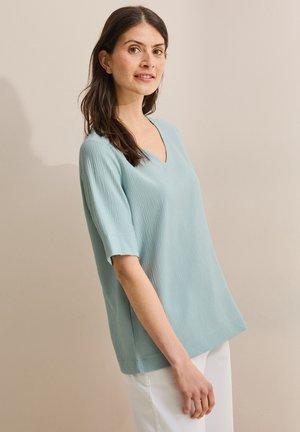 Haut texturé bleu clair avec un décolleté en V et des manches courtes. Design à coupe droite et détails de couture subtils. Associé à un pantalon blanc.