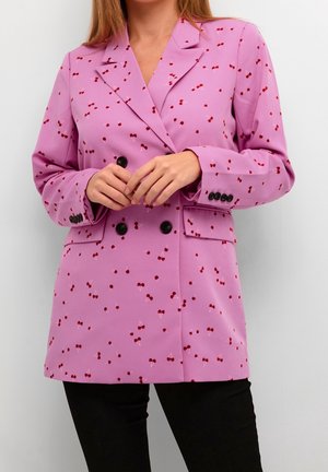 Femme portant un blazer rose à double boutonnage avec des motifs de cerises rouges, des boutons noirs et un pantalon noir, mains jointes devant.