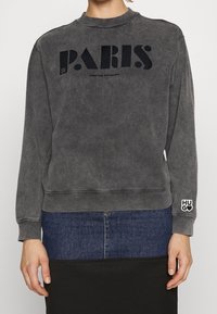 Sudadera gris desgastada con escote redondo, gráfico "PARIS" en negro, puños acanalados y logo "HUGO" en la manga. Tejido suave y texturizado.