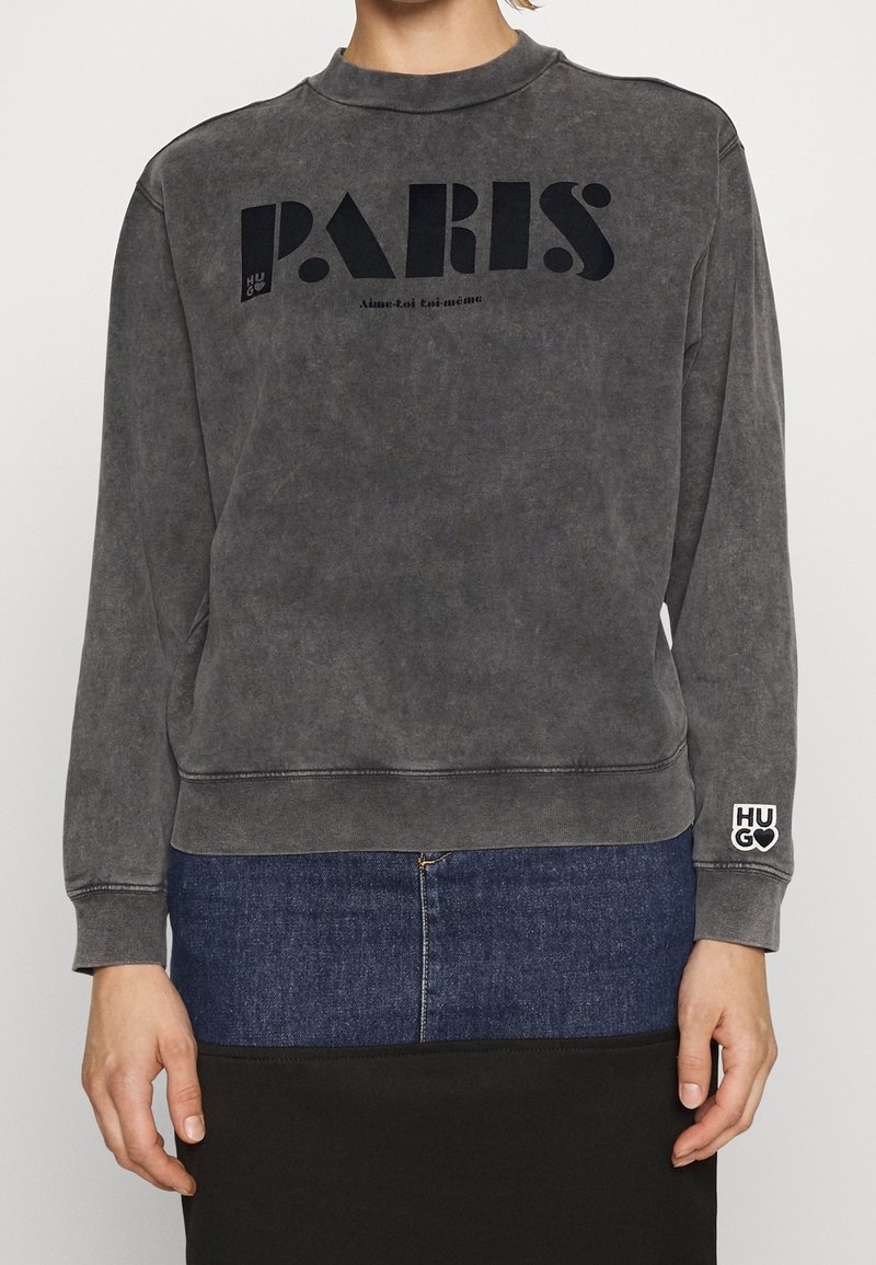 Sudadera gris desgastada con escote redondo, gráfico "PARIS" en negro, puños acanalados y logo "HUGO" en la manga. Tejido suave y texturizado.