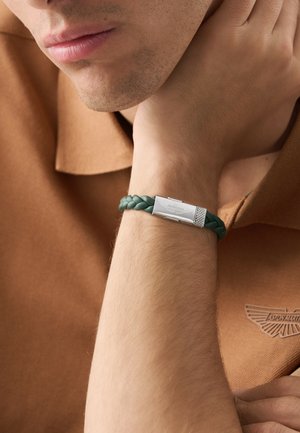 Homme portant un bracelet en cuir tressé vert avec un fermoir en argent gravé d'un logo, vêtu d'une chemise marron avec un logo assorti.
