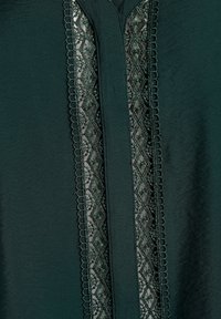 Tessuto verde scuro con un pannello verticali centrale in pizzo che presenta motivi a diamante e floreali, bordato da un orlo a scallop.
