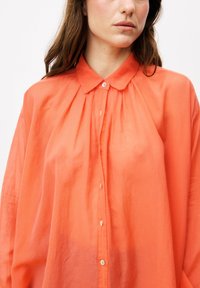 Korallenfarbene Hemd mit Knopfleiste, das einen Kragen, eine gefältelte Vorderseite und einen leichten Stoff aufweist. Bemerkenswerte Details sind Knöpfe aus Perlmutt und eine lockere Passform.