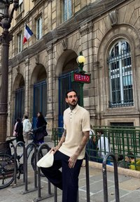 Homme avec des tatouages et un polo beige tenant un chapeau blanc près de l'entrée du métro de Paris dans une rue de la ville avec un bâtiment historique et des vélos.