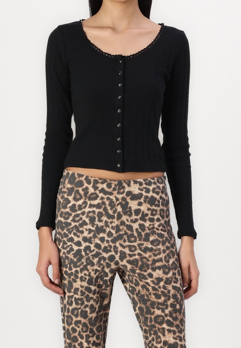 Femme portant un crop top noir à manches longues boutonné associé à un pantalon taille haute imprimé léopard, debout devant un fond uni.