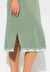 Abito in cotone verde con uno spacco laterale e un bordo in pizzo scallopato di tonalità più chiara all'orlo, che conferisce un tocco decorativo sottile.