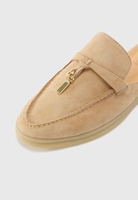 Beige suede slip-on shoe featuring a rounded toe, a small bow detail, and a gold-tone accent near the top. Soft texture with minimal stitching.