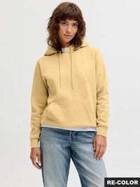 Jonge vrouw met krullend bruin haar, een beige hoodie en een blauwe spijkerbroek, staand tegen een effen lichtgrijze achtergrond.