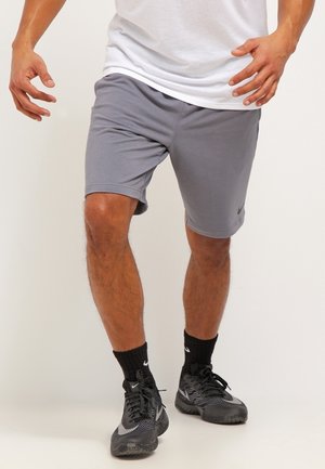 Homme portant un short de sport gris, des chaussettes noires et des baskets Nike noires et grises, debout avec les jambes légèrement pliées et les bras levés.