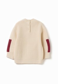 Beige gestrickter Pullover mit geripptem Design, drei Knöpfen auf der Rückseite und burgundy Akzentpaneelen an den Ärmeln und der Tasche.