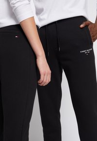 Black sweatpants with drawstring waist, side pockets, and embroidered "Tommy Hilfiger" logo in white and red accents. Smooth fabric texture.