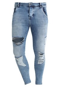 Jeans de denim azul claro con parches desgastados, que presentan múltiples desgarros y bordes deshilachados. Diseño ajustado con cierre tradicional de botón y cremallera.