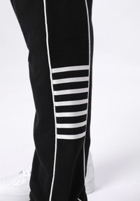 Zwarte trainingsbroek met witte zijstrepen. De onderkant van de broekspijp heeft een reeks witte horizontale strepen. Gemaakt van zachte, gestructureerde stof.