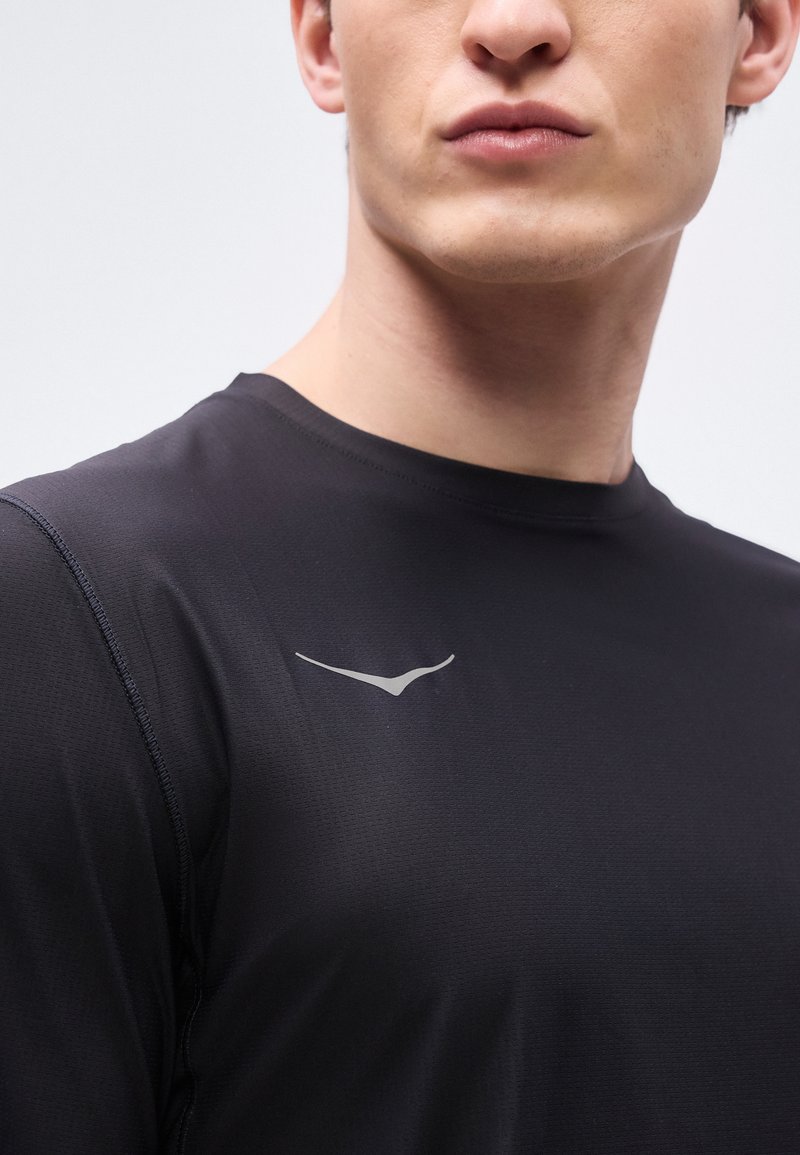 Primo piano di una persona che indossa una maglietta sportiva nera con un piccolo logo riflettente vicino alla spalla sinistra su uno sfondo uniforme.