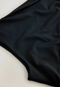 Fekete, sima anyagú fürdőruha alsó, ívelt szélekkel, egy világos felületen kiterítve.