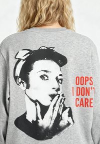 Personne blonde portant un sweat gris avec un visage de femme en noir et blanc qui envoie un baiser, avec un texte rouge « OOPS I DON’T CARE » dans le dos.