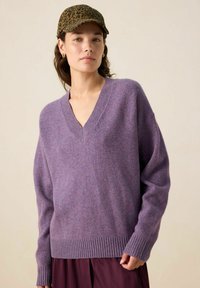 Un pull-over violet en v fait d'un matériau en tricot doux avec un ourlet et des poignets côtelés ; porté avec une casquette imprimée léopard.