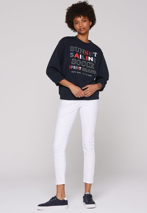 Marineblaues Sweatshirt mit grafischem Text in Rot, Weiß und Blau, kombiniert mit weißen Skinny-Jeans und dunklen Sneakers. Verfügt über eine lockere Passform und lange Ärmel.