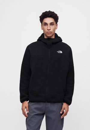 Мъж, облечен в черно флийс яке с цип и качулка с логото на The North Face на гърдите, стои срещу светъл фон.
