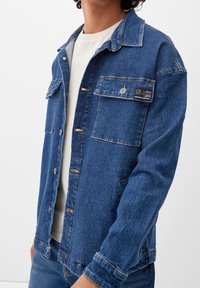 Chaqueta de mezclilla en azul oscuro con bolsillos frontales, cierre de botones y costuras de contraste. Llevada sobre una camisa de color claro.