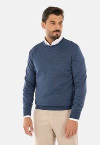 Maglione di lana blu a maglia con scollatura rotonda e maniche lunghe, caratterizzato da un orlo e polsini a costine, indossato sopra una camicia bianca con colletto.