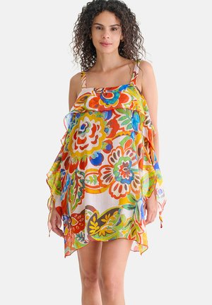Femme aux cheveux bouclés portant une robe fleurie ample et colorée avec des motifs vifs orange, jaune, vert et bleu sur fond blanc.