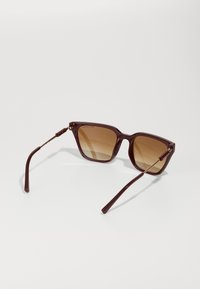 Ochelari de soare cu nuanță maronie și cadru din plastic cărămiziu. Formă pătrată cu accente metalice subțiri pe tâmple. Textură netedă, design ușor.