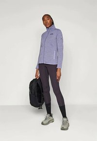 Φούτερ σε ανοιχτό μωβ χρώμα με ψηλό γιακά, φερμουάρ στον μπροστινό μέρος και πλαϊνές τσέπες, συνδυασμένο με μαύρα κολάν και γκρι παπούτσια, κρατώντας ένα μαύρο σακίδιο.