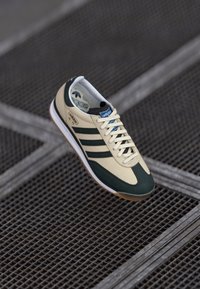 Beige sneaker met groene accenten en drie donkergroene strepen, met een ronde neus, gestructureerde bovenkant en een gumrubberen zool.