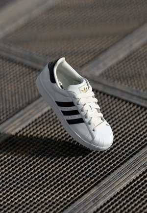 White sneaker with a textured rubber sole, black stripes, and a black heel tab. Features a rounded toe and lace-up front with gold logo detail.