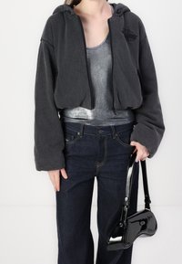 Sudadera negra recortada con capucha, combinada con una camiseta sin mangas plateada metálica y jeans de pierna ancha azul oscuro. Lleva un bolso de mano de charol negro.