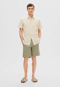 Lys beige kortermet skjorte, grønne shorts, lyse sandaler. Avslappet passform med knappdetaljer, glatt tekstur og enkelt design.