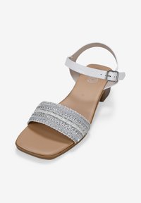 Sandale blanche à lanières avec un design à bout carré, ornée de brides en argent et d'un petit talon carré. Semelle intérieure en beige lisse. Boucle de cheville réglable.