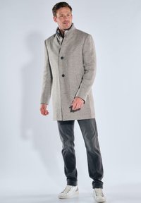 Hellgrauer Wollmischmantel mit hohem Kragen, ausgestattet mit drei Knöpfen. Getragen über dunklen Jeans und weißen Turnschuhen. Glatte Textur.