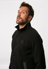 Schwarze Zip-Jacke mit hohem Kragen, weichem Material und einem Logo-Detail auf der Vorderseite. Sie verfügt über kontrastierende graue Reißverschlussakzente an den Taschen.