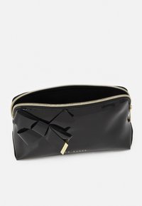 Svart lackläderpouch med gulddragkedja, en rosettdetalj på framsidan och branding på botten. Slät textur och strukturerad form.