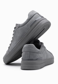 Zapatillas de ante gris con acabado texturizado, suela de goma plana y acento metálico. Presentan un diseño con cordones y detalles de logo en el lateral.