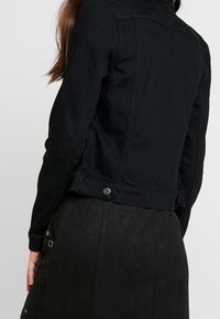 Woman wearing a fitted black denim jacket with button details, shown from the back, alongside a black skirt with metal eyelets.