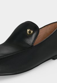 Mocassin en cuir noir avec une petite décoration métallique dorée en forme de cœur sur le dessus, doublure intérieure beige et petit talon.