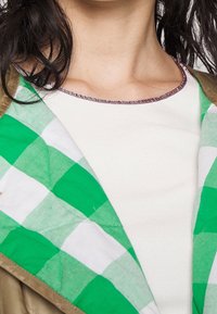 Femme portant un pull côtelé blanc sous un manteau avec une doublure à carreaux verts et blancs, montrant le décolleté et des mèches de cheveux partiellement visibles.