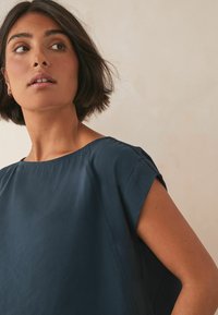 Granatowa bluzka z krótkim rękawem o szerokim dekolcie i przeszyciami, wykonana z lekkiego materiału, charakteryzująca się luźnym krojem i gładką teksturą.